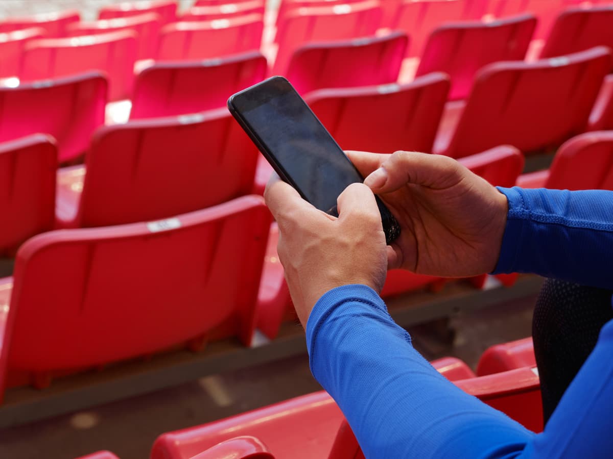 Personas sēž stadionā, tur mobilu tālruni un skatās uz ekranu, apstāda ar tukšām vietām