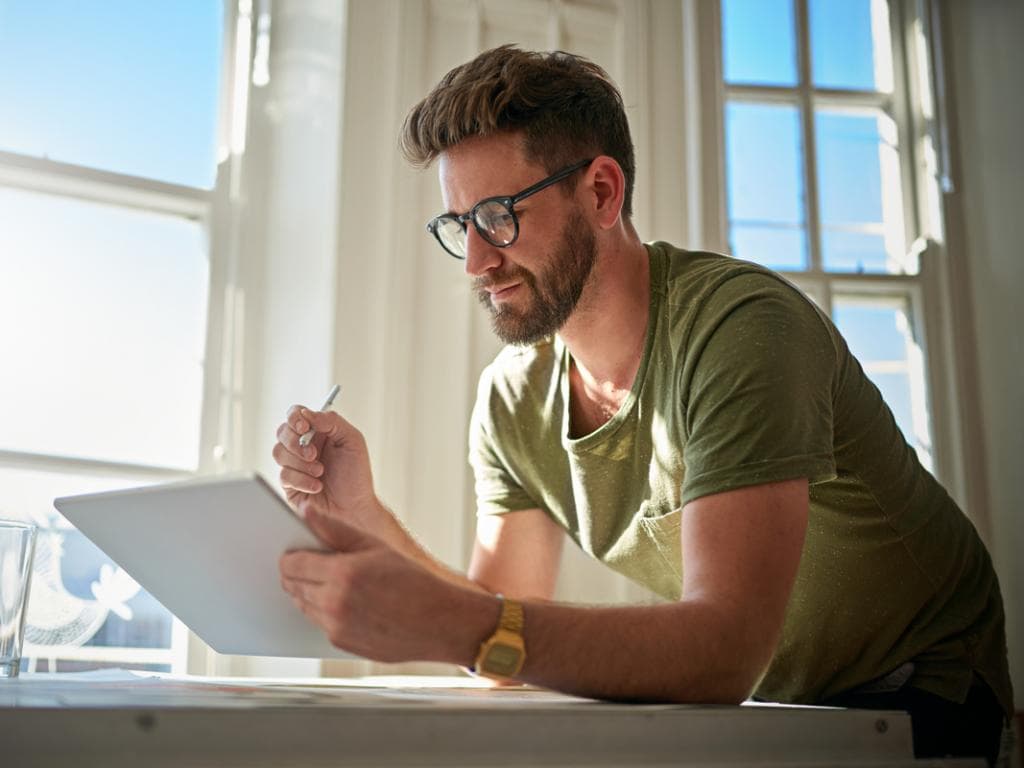 Vīrietis ar brillēm sēž pie galda, koncentrējoties uz planšetdatoru priekšā