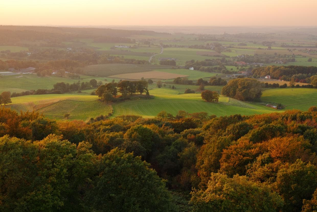 Skats uz zaļu ieleju, ko ieskauj koki un viļņaini kalni zem dzidri zilas debess