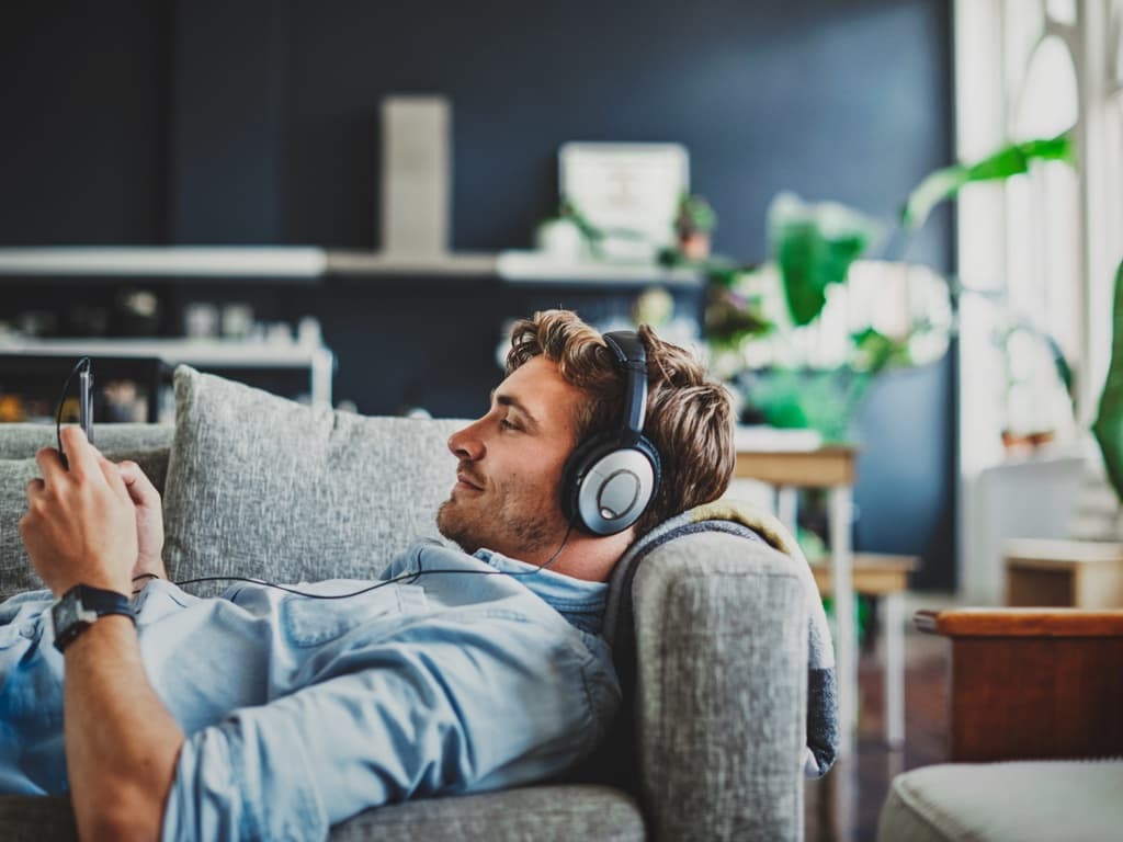 Vīrietis sēž uz dīvāna ar austiņām, koncentrējoties uz tālruņa ekrānu