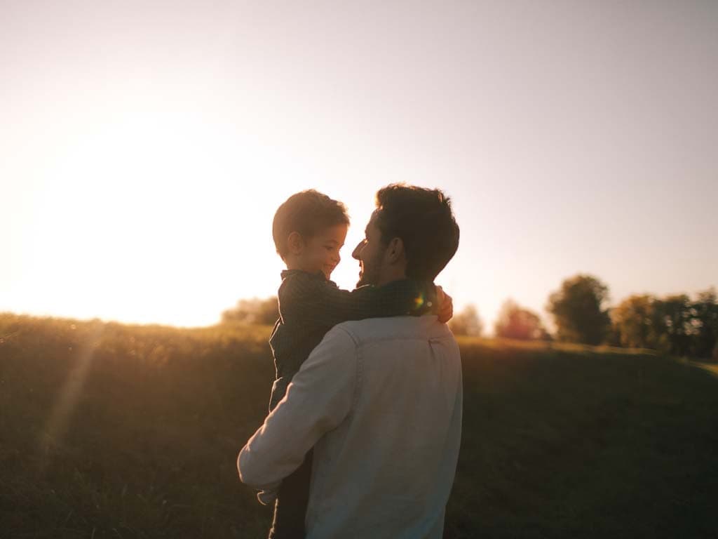 Tēvs un dēls stāv kopā laukā saulrietā, siltiem toņiem izgaismojot debesis aiz viņiem