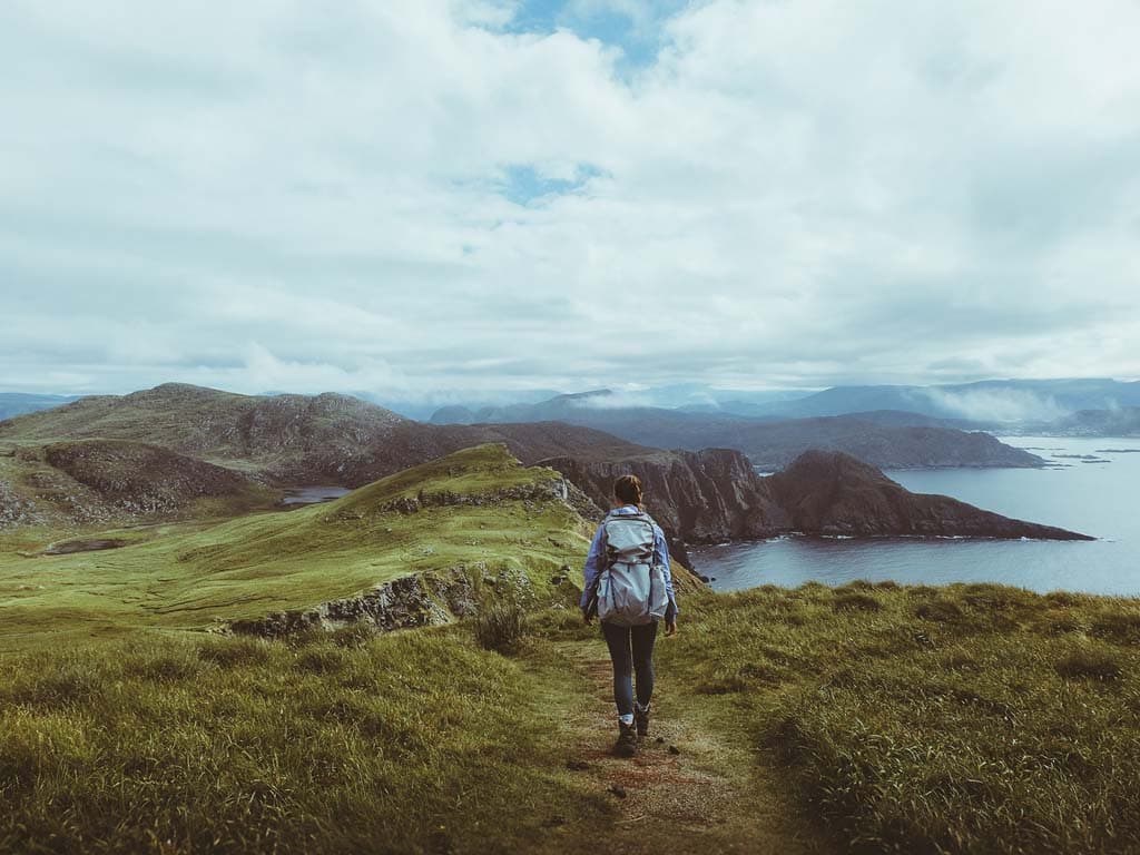 Persona pastaigājas pa gleznainu taku ar skatu uz okeānu un kalniem fonā
