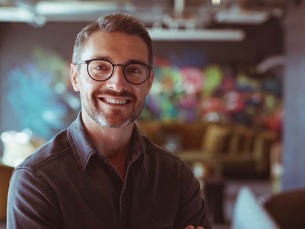 Vīrietis ar brillēm un melnu kreklu sirsnīgi smaida kamerai