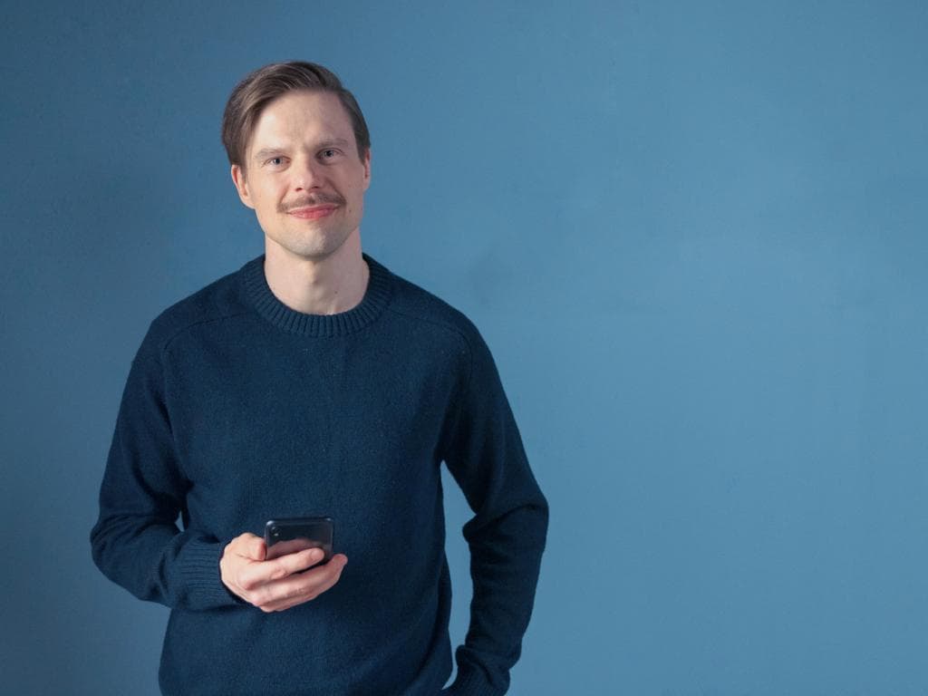 Vīrietis džemperī tur labajā rokā mobilo telefonu, skatoties uz ekrānu ar koncentrētu izteiksmi