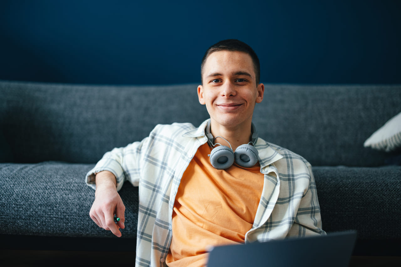 Jauns vīrietis ar austiņām sēž uz dīvāna, koncentrējies uz savu klēpjdatoru mājīgā viesistabas vidē